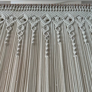 May include: A white macrame wall hanging with a decorative pattern at the top. The hanging is made of a thick, natural fiber rope.