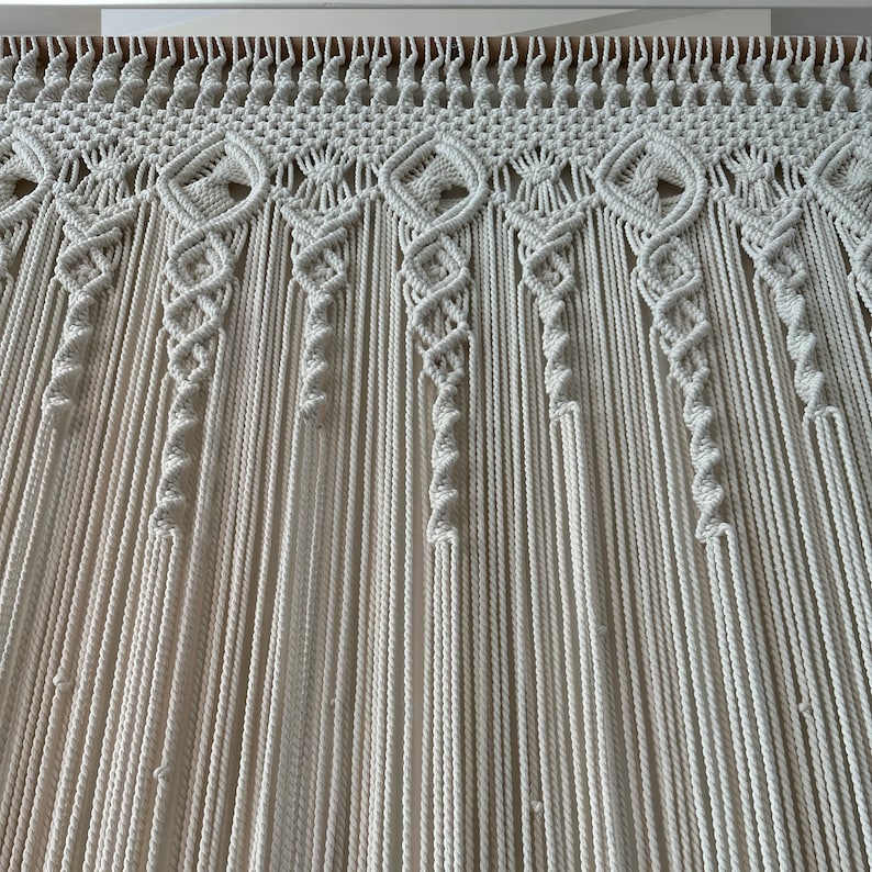 May include: A white macrame wall hanging with a decorative pattern at the top. The hanging is made of a thick, natural fiber rope.