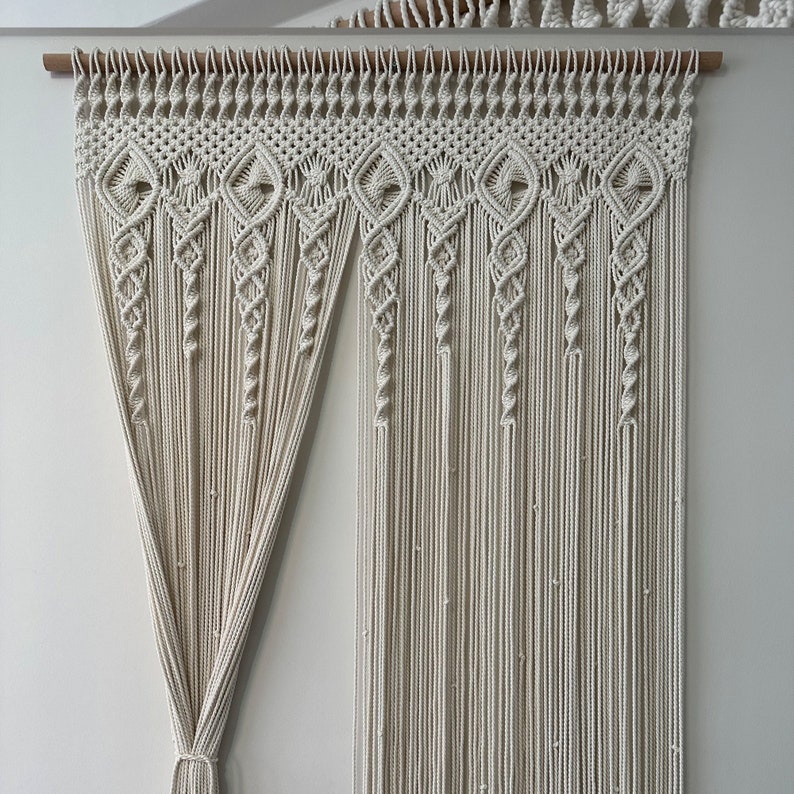 May include: A white macrame wall hanging with a geometric pattern. The hanging is made of cotton cord and is attached to a wooden dowel.