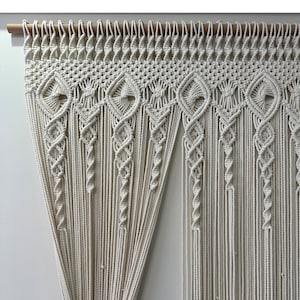 May include: A white macrame wall hanging with a geometric pattern. The hanging is made of cotton cord and is attached to a wooden dowel.