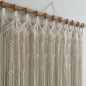 May include: A white macrame wall hanging with a wood dowel rod and brown hooks. The macrame is made with a knot pattern that creates a series of arches.