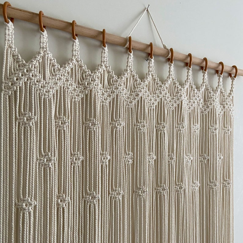 May include: A white macrame wall hanging with a wood dowel rod and brown hooks. The macrame is made with a knot pattern that creates a series of arches.
