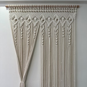 May include: A white macrame wall hanging with a decorative pattern. The hanging is made of cotton cord and is attached to a wooden dowel rod.