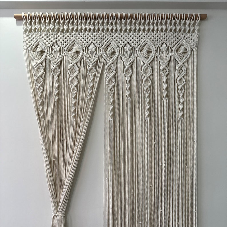 May include: A white macrame wall hanging with a decorative pattern. The hanging is made of cotton cord and is attached to a wooden dowel rod.