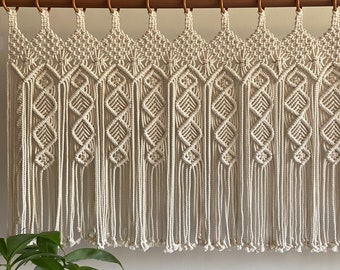May include: A white macrame wall hanging with a geometric pattern and fringe. The hanging is attached to a wooden dowel rod with rings.