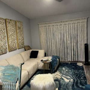 May include: A living room with a white macrame wall hanging, a white sectional sofa, a blue and white rug, a white faux fur ottoman, and a gold side table.