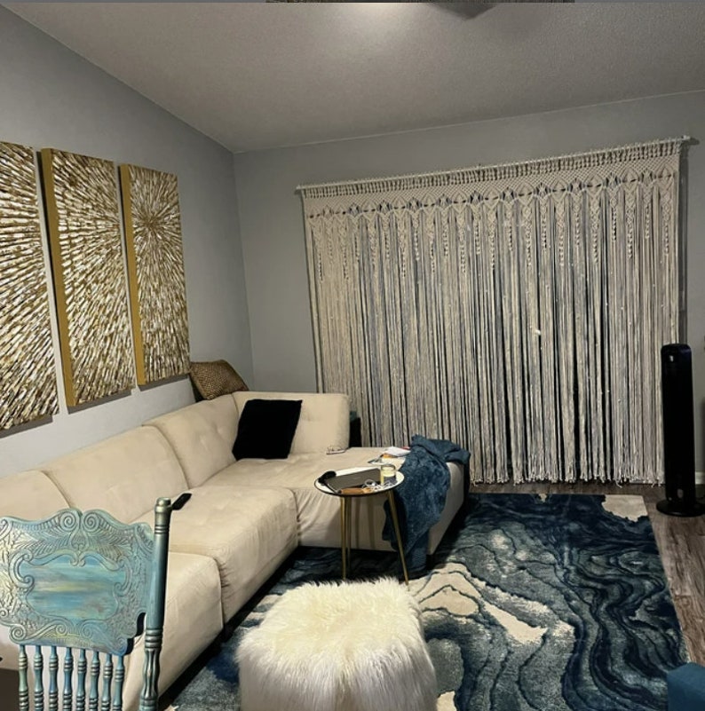 May include: A living room with a white macrame wall hanging, a white sectional sofa, a blue and white rug, a white faux fur ottoman, and a gold side table.