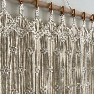May include: A white macrame wall hanging with a geometric pattern. The hanging is made of cotton cord and is attached to a wooden dowel rod.