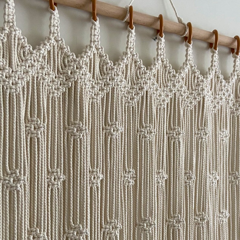 May include: A white macrame wall hanging with a geometric pattern. The hanging is made of cotton cord and is attached to a wooden dowel rod.