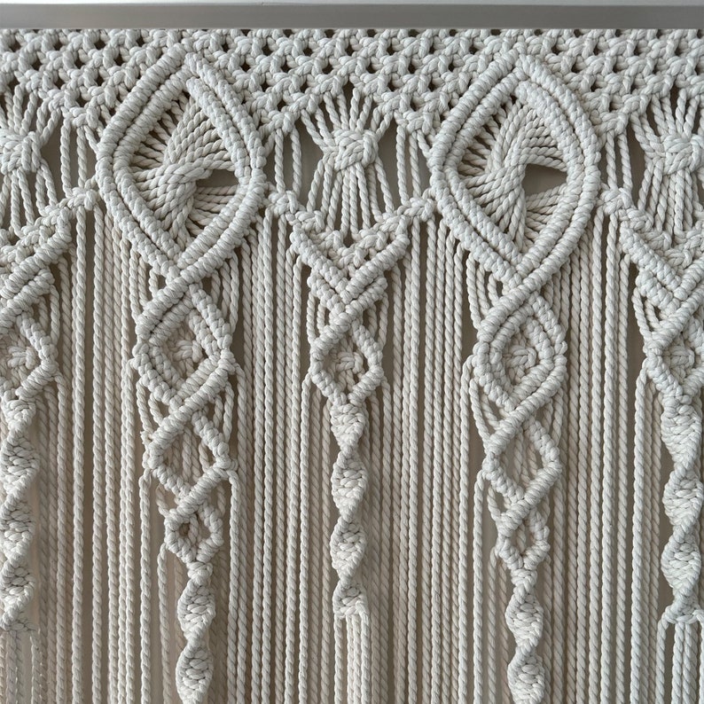 May include: A white macrame wall hanging with a geometric pattern. The hanging is made of thick cotton cord and has a fringe at the bottom.