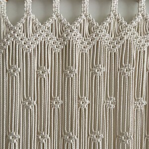 May include: A white macrame wall hanging with a geometric pattern. The hanging is made of thick cotton cord and has a fringe at the bottom.
