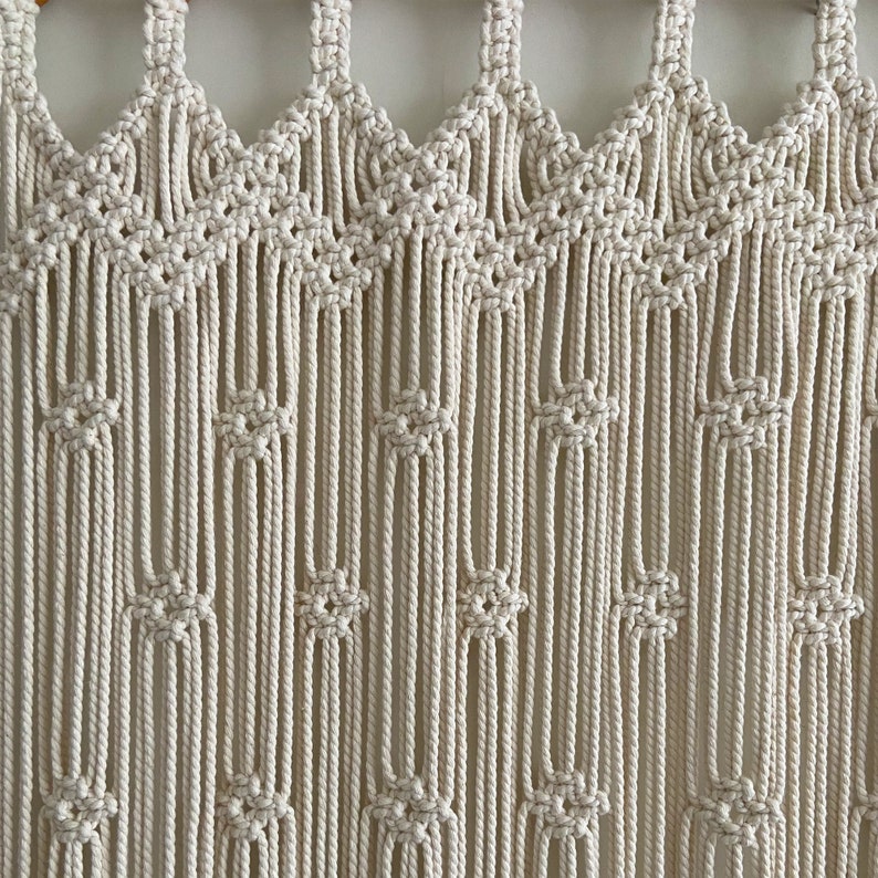May include: A white macrame wall hanging with a geometric pattern. The hanging is made of thick cotton cord and has a fringe at the bottom.
