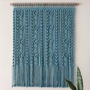 Op de afbeelding: Een tealblauwe macramé wandhanger met een geometrisch patroon. De hanger heeft een houten lat aan de bovenkant en lange franjes aan de onderkant.