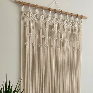 May include: A white macrame wall hanging with a geometric pattern. The hanging is made of natural cotton cord and is attached to a wooden dowel rod.