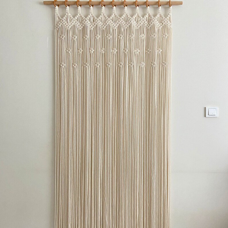May include: A white macrame wall hanging with a geometric pattern. The hanging is made of cotton cord and is attached to a wooden dowel rod.