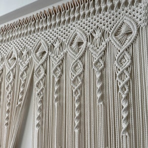 May include: A white macrame wall hanging with a geometric pattern. The hanging is made of thick cotton cord and has a wooden dowel at the top.