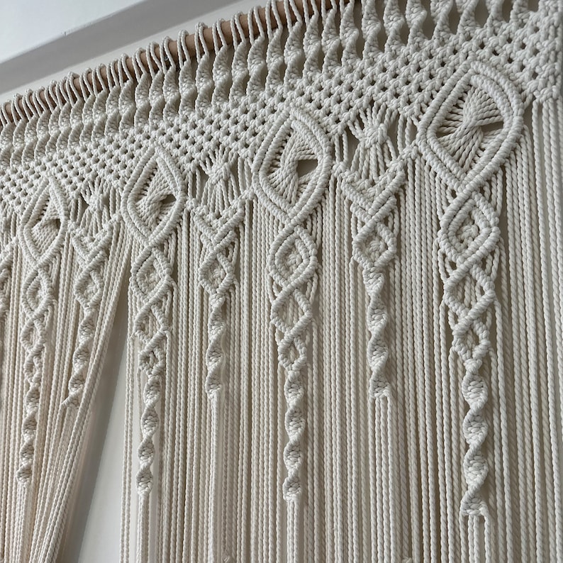 May include: A white macrame wall hanging with a geometric pattern. The hanging is made of thick cotton cord and has a wooden dowel at the top.