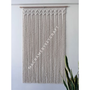 May include: A white macrame wall hanging with a geometric pattern at the top and beads throughout. The hanging is made of natural cotton cord and is approximately 90 cm wide and 150 cm tall.