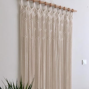 May include: A white macrame wall hanging with a geometric pattern. The hanging is made of cotton cord and is attached to a wooden dowel rod.