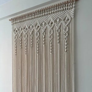 May include: A white macrame wall hanging with a geometric pattern. The hanging is made of cotton cord and is attached to a wooden dowel.
