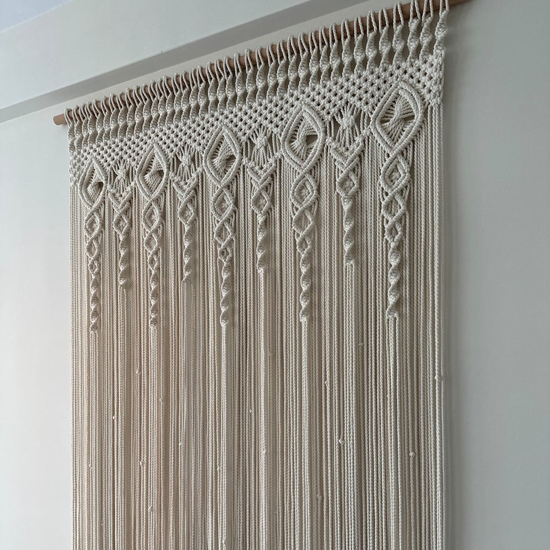 May include: A white macrame wall hanging with a geometric pattern. The hanging is made of cotton cord and is attached to a wooden dowel.