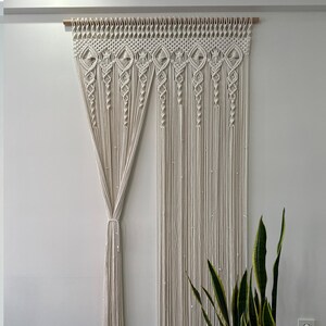 May include: A white macrame curtain with a geometric pattern hangs from a wooden rod. The curtain is made of thick cotton cord and has a fringed bottom.