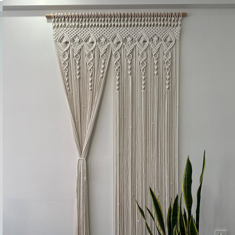 May include: A white macrame curtain with a geometric pattern hangs from a wooden rod. The curtain is made of thick cotton cord and has a fringed bottom.