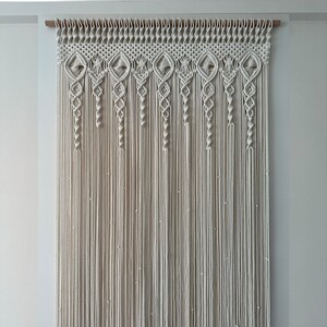 May include: A white macrame wall hanging with a geometric pattern. The hanging is made of cotton cord and is attached to a wooden dowel.