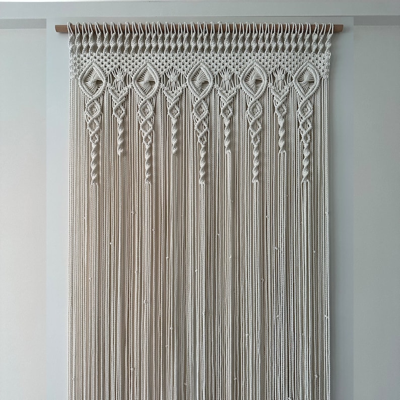 May include: A white macrame wall hanging with a geometric pattern. The hanging is made of cotton cord and is attached to a wooden dowel.