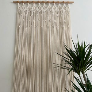 May include: A white macrame wall hanging with a geometric pattern. The hanging is made of natural cotton cord and is attached to a wooden dowel rod.