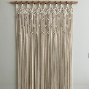 May include: A white macrame wall hanging with a geometric pattern. The hanging is made of knotted cords and is attached to a wooden dowel rod.