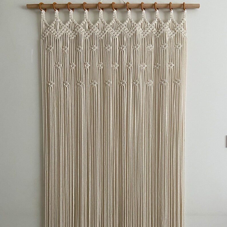May include: A white macrame wall hanging with a geometric pattern. The hanging is made of knotted cords and is attached to a wooden dowel rod.