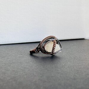 May include: A white gemstone ring with a copper wire design. The ring is set on a dark gray surface.