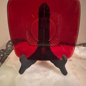 May include: A square, translucent red glass plate featuring a central raised vertical pillar design with a circular base and radiating linear patterns. The plate is displayed on a black plastic stand, showcasing its deep crimson hue and smooth finish.