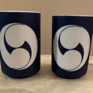 May include: Two cylindrical cups with a matte dark blue exterior and a white interior. Each cup features a large, centered white circular emblem containing a stylized, three-part swirling design in dark blue, creating a bold contrast.