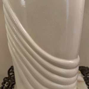 Puede incluir: Un jarrón de cerámica de color blanco roto brillante que presenta una serie de crestas diagonales en relieve que envuelven la parte inferior izquierda y la base de la estructura, creando un aspecto texturizado y superpuesto.