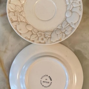 May include: Two off-white ceramic saucers. The top saucer features a raised relief pattern of fruits and flowers along the rim. The bottom saucer is flipped over, revealing a stamp that reads Vernon Ware by Metlox Made in California.