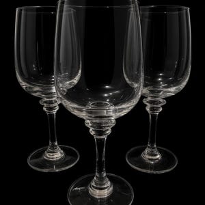 May include: A set of three clear glass wine glasses arranged in a triangular formation. Each glass features a smooth, rounded bowl, a slender stem with a decorative ring detail near the top, and a circular base, all set against a solid black background.