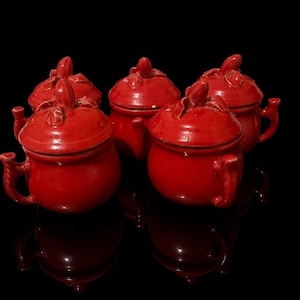 May include: A set of four small, glossy red ceramic pots with lids. Each pot features a rounded body, a single side handle, and a lid topped with a sculpted leaf and stem detail. The pots have a smooth, reflective finish and uniform color.