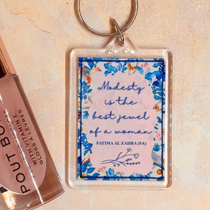 Puede incluir: Llavero acrílico transparente con un diseño floral y la cita "La modestia es la mejor joya de una mujer" de Fátima Al Zahra (SA). El llavero tiene un anillo de metal plateado.