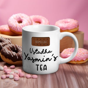 Peut inclure: Une tasse en céramique blanche avec un motif de biscuit brun et le texte "Bismillah" dessus. La tasse a également un texte noir qui dit "Ustadha Yasmin's TEA".