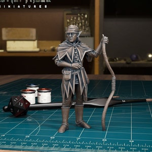 May include: A painted miniature figurine of an elf wearing a hooded cloak and holding a staff. The figurine is standing on a green cutting mat with a ruler and paint pots.