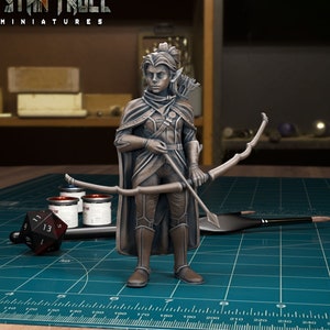 May include: A painted miniature figurine of an elf archer, holding a bow and arrow. The elf is wearing a brown leather tunic, trousers, and boots. The miniature is painted in a metallic bronze colour. The miniature is on a green cutting mat with a ruler and paint pots.