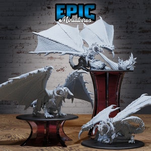 Può includere: Tre miniature di draghi dipinte di bianco su stand espositivi. I draghi sono in varie pose, con le ali spiegate e le code arrotolate. Gli stand sono di colore rosso scuro. L'immagine include il testo "EPIC Miniatures".
