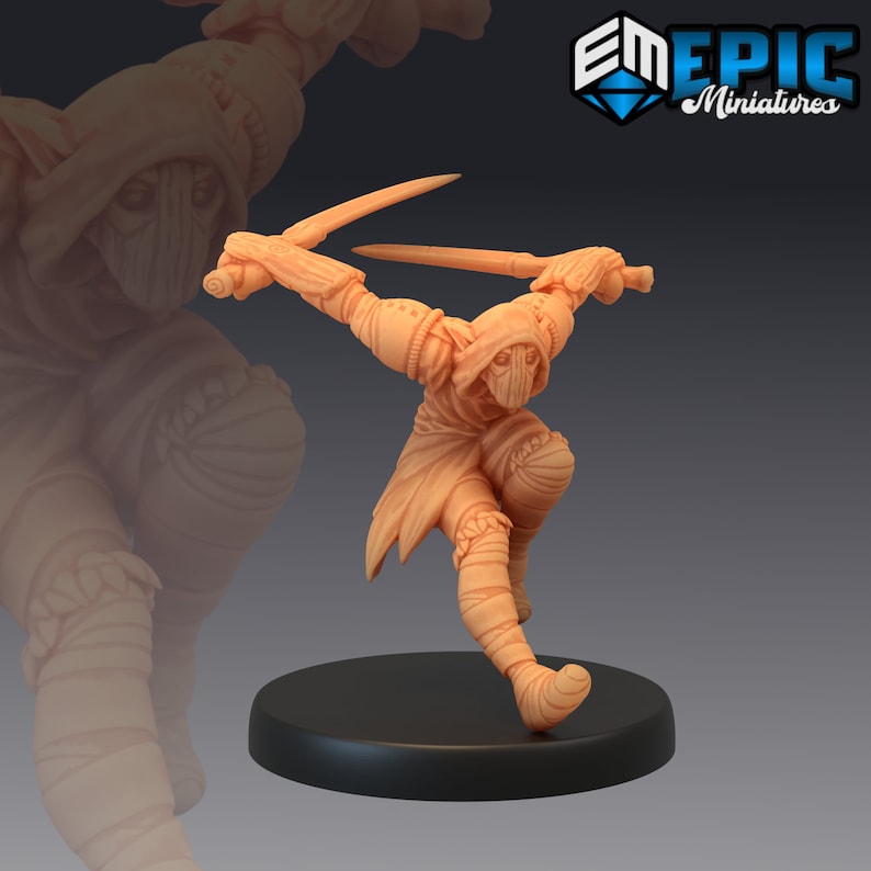 May include: A miniature figurine of a hooded figure in a fighting pose, holding two swords. The figure is wearing a hooded cloak and has a mask over their face. The figure is painted in a light brown colour.