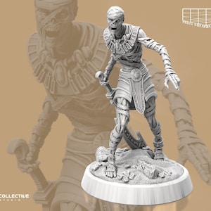 Könnte beinhalten: Eine weiß bemalte Miniaturfigur einer Mumie mit Bandagen, die ein Krummschwert hält. Die Mumie steht auf einem runden Sockel mit Sandtextur.