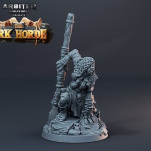 Op de afbeelding: Een grijze plastic miniatuurfiguur van een Ork-krijger die een staf vasthoudt. De Ork draagt een capuchon en heeft een woeste uitdrukking op zijn gezicht. De miniatuur staat op een rotsachtige basis.