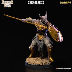 Könnte beinhalten: Eine bemalte Miniaturfigur eines Serpopards, einer Kreatur mit einem Schakal-Kopf und einem menschlichen Körper, die eine goldene Rüstung trägt und einen Speer und einen Schild hält. Die Figur steht auf einem felsigen Sockel.