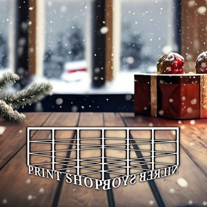 以下が含まれることがあります： 黒い文字で「PRINT SHOP」と書かれた白い木製のフレーム。フレームは赤いギフトボックスとクリスマスツリーの枝を背景に、木製のテーブルの上に置かれています。シーンは軽い雪で覆われています。