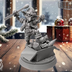 Puede incluir: Una figura en miniatura gris de un duende sosteniendo una cerilla grande sobre una caja de regalos. La figura está sobre una base redonda. El fondo incluye un árbol de Navidad y regalos envueltos.